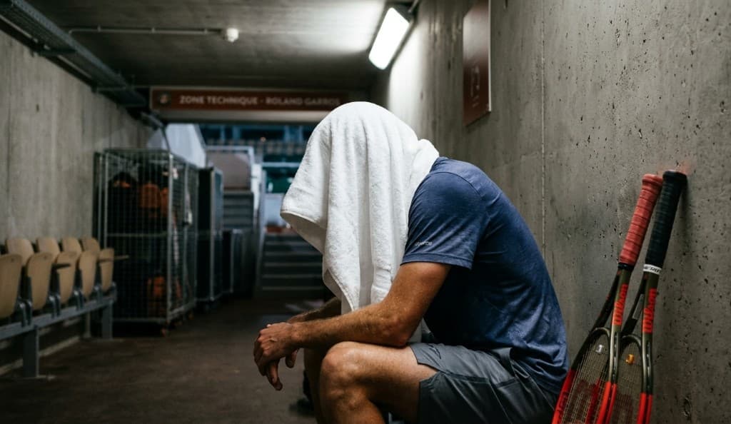 A tennis player sits exhausted in a Roland Garros tunnel with a towel over his head and Wilson rackets leaning against the concrete wall